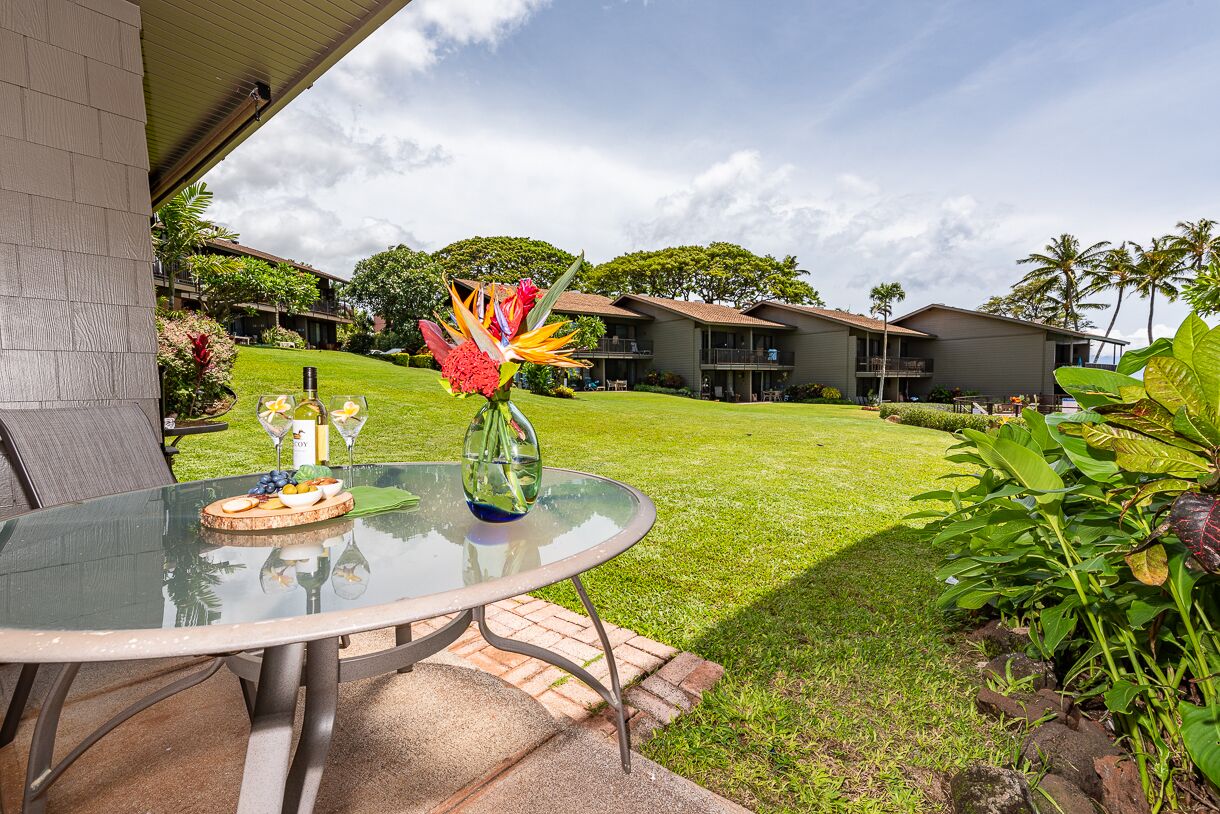 A glass table with a fruit platter, a bottle, and a vase with flowers, set on a patio with a view of a well-maintained lawn and multiple buildings in the background.