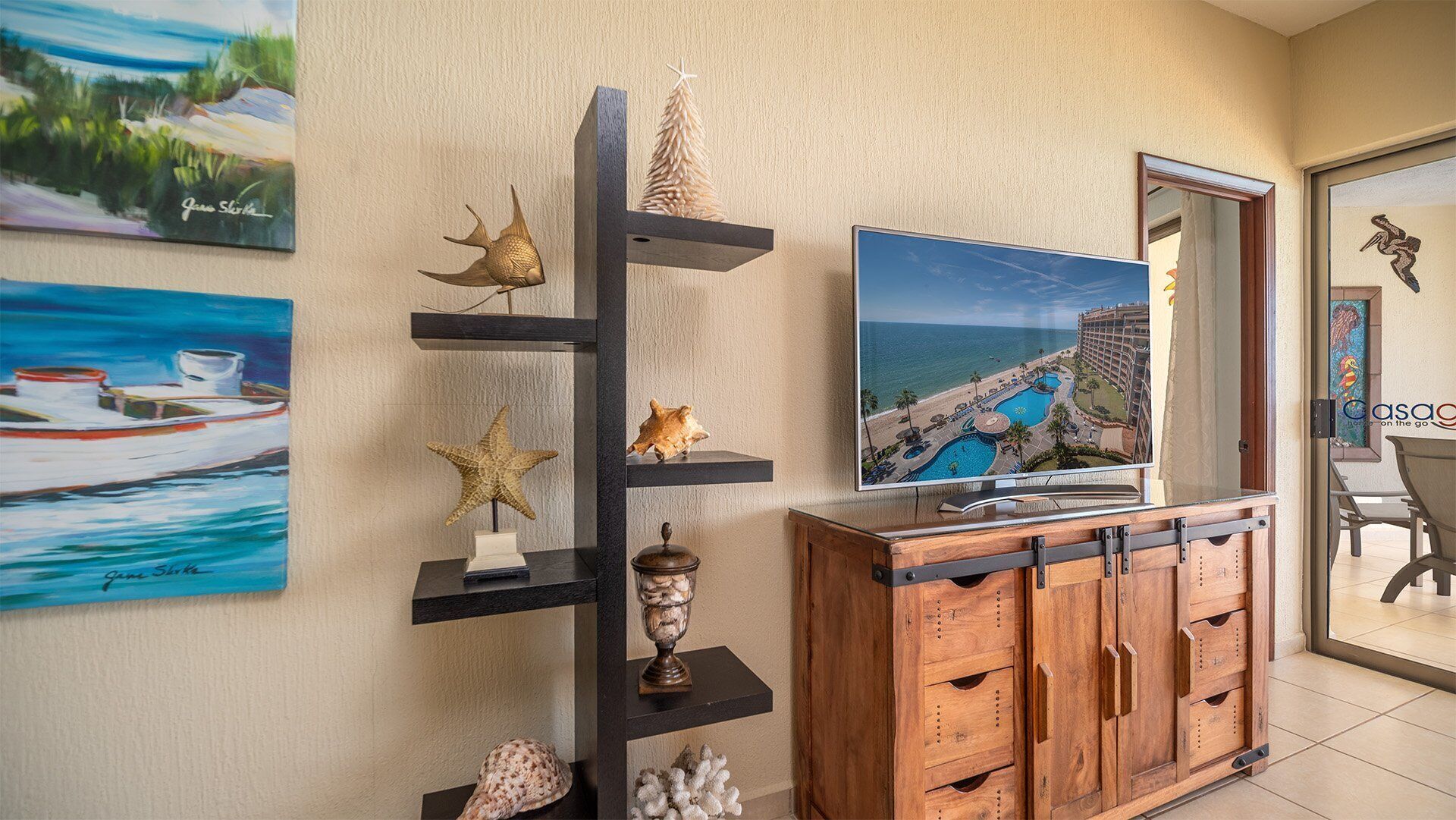 Ein Zimmer mit Fernseher, der einen Blick auf ein Resort zeigt, einem Holzkasten und Regalen mit dekorativen Muscheln und einem Seestern.