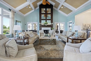 A spacious living room with a high ceiling, large windows, and a fireplace.