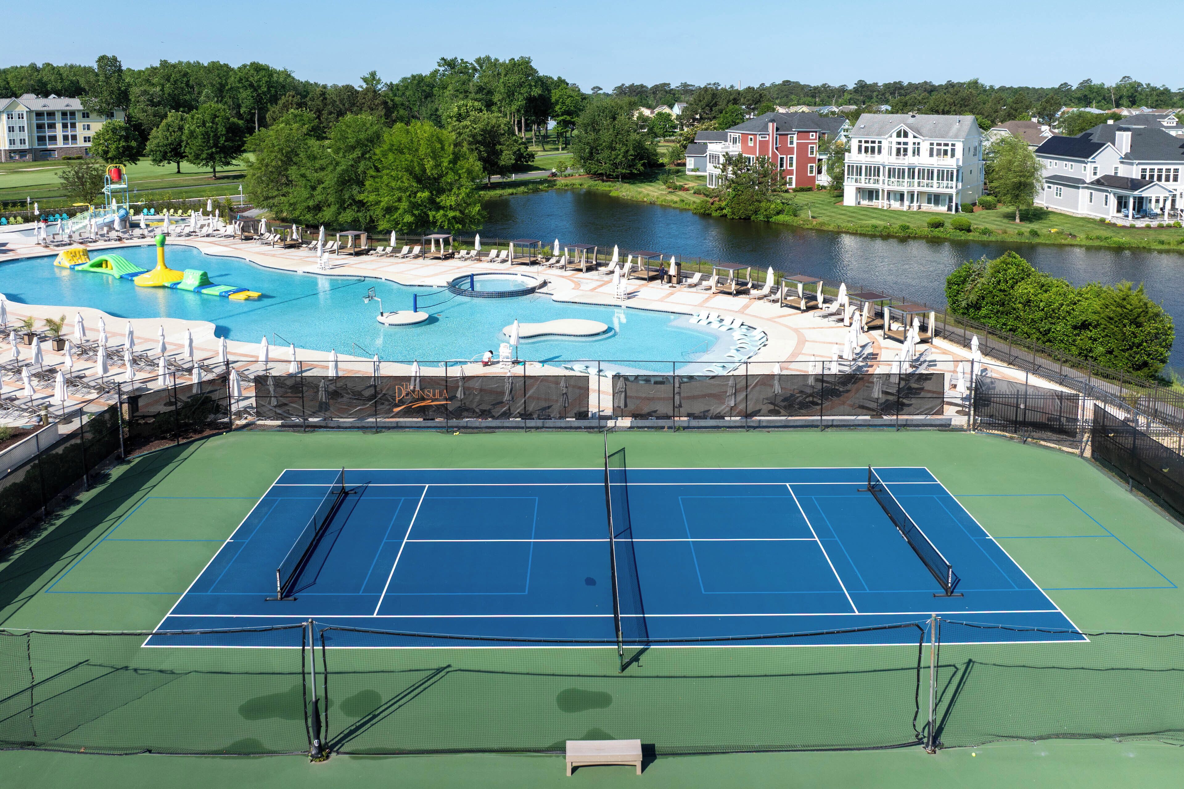 Una cancha de tenis con red, rodeada por una zona de piscina con sillones de descanso y un tobogán.