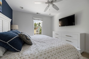 A bedroom with a bed, a dresser, a ceiling fan, a window, and a TV mounted on the wall.