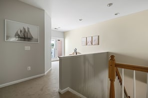 A hallway with a white door leading to a balcony, a framed picture of sailboats on the left wall, and a wooden railing on the right.