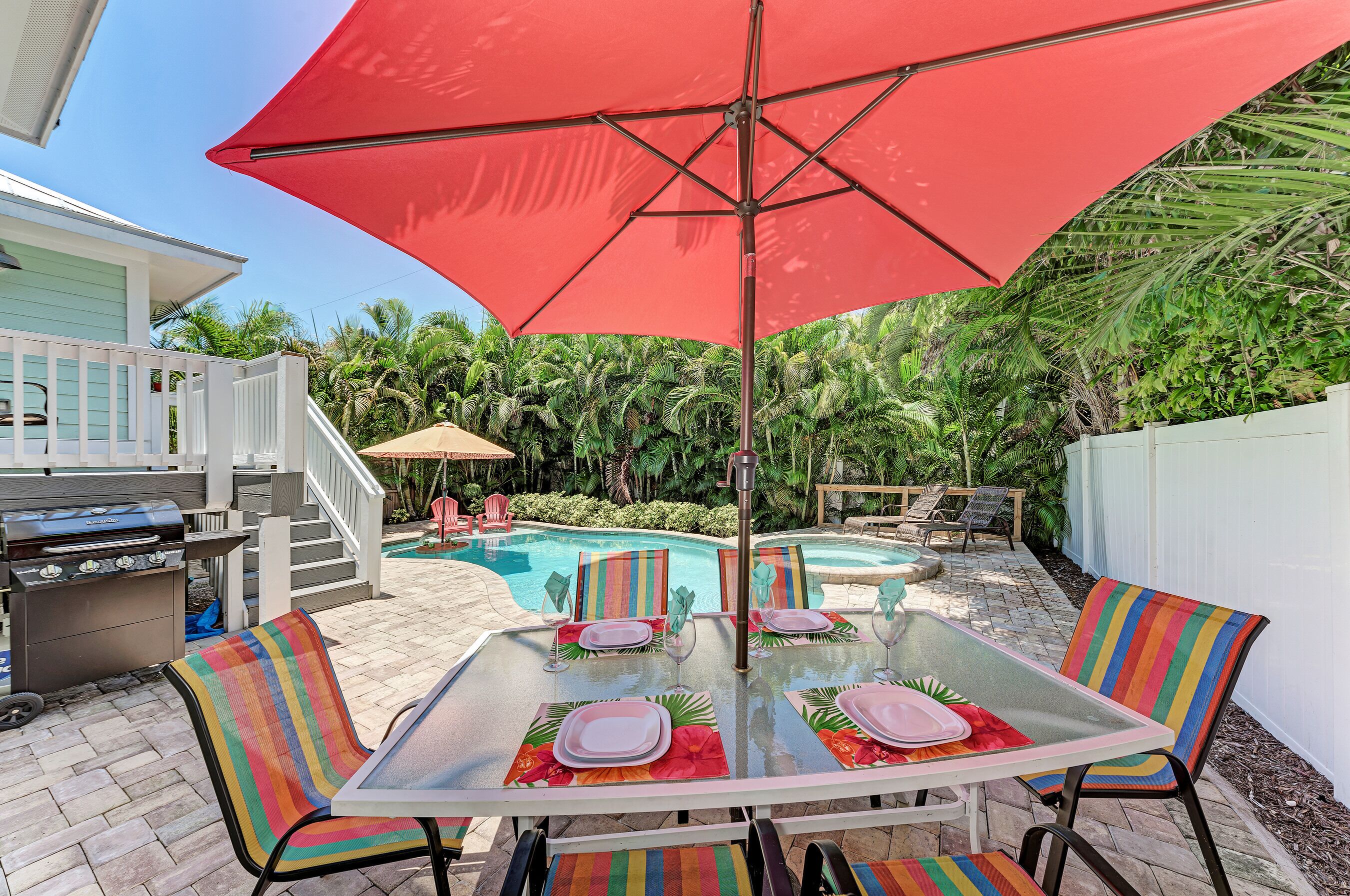 A backyard with a glass table, colorful striped chairs, a red umbrella, and a pool.