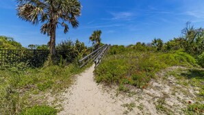 A sandy path leading to a wooden bridge, surrounded by greenery and palm trees under a clear blue sky. - Modern Beach Retreat ~ Close to the Beach! (Saint Augustine)
