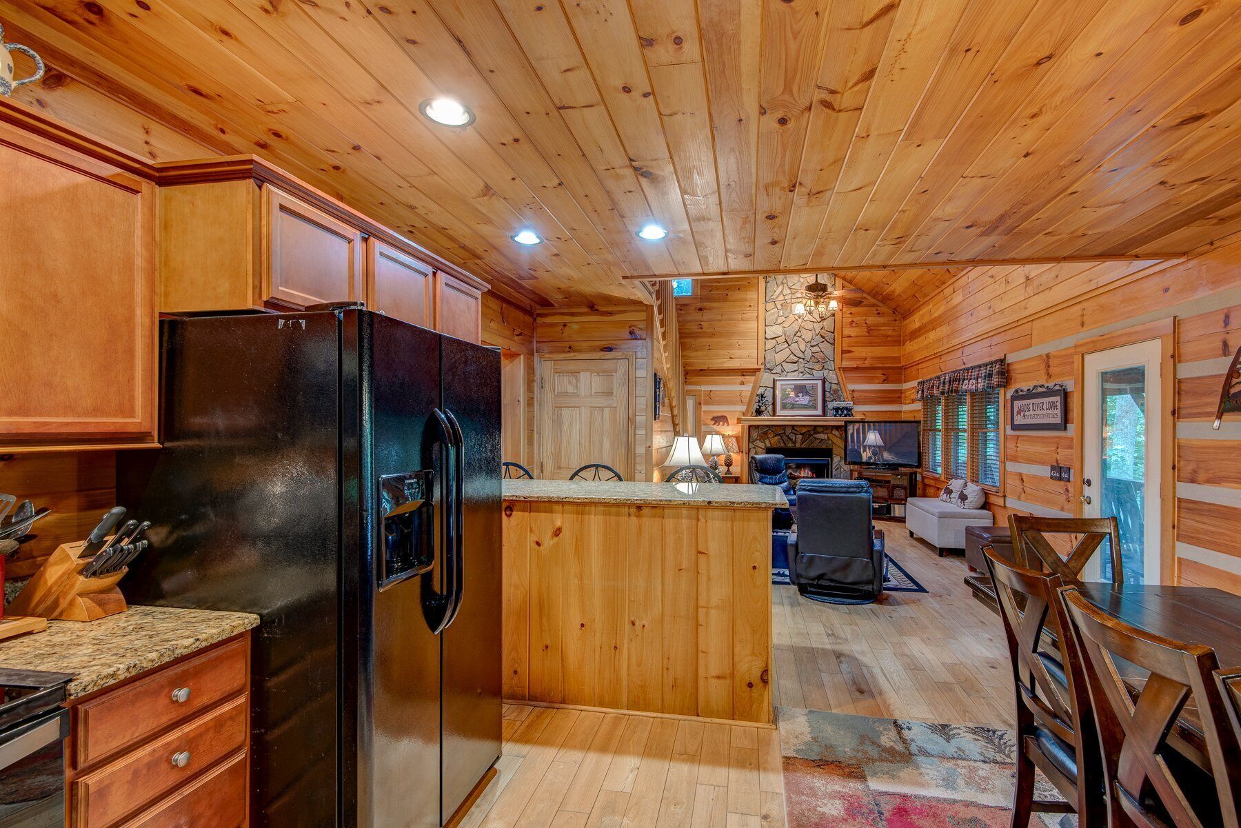 A cabin interior with a kitchen area featuring a black refrigerator, wooden cabinets, and a stone fireplace.