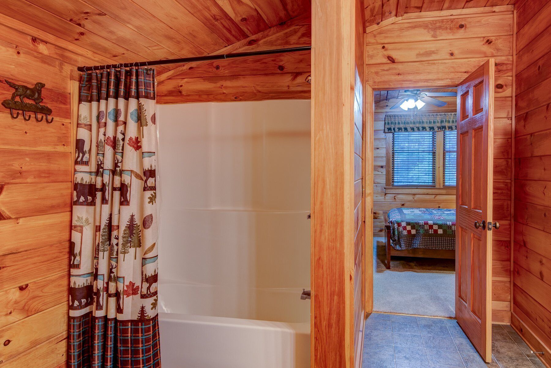 A wooden cabin interior with a visible bathroom, a bedroom with a bed, and a window with blinds.