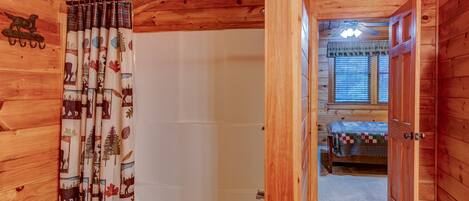 A wooden cabin interior with a visible bathroom, a bedroom with a bed, and a window with blinds.