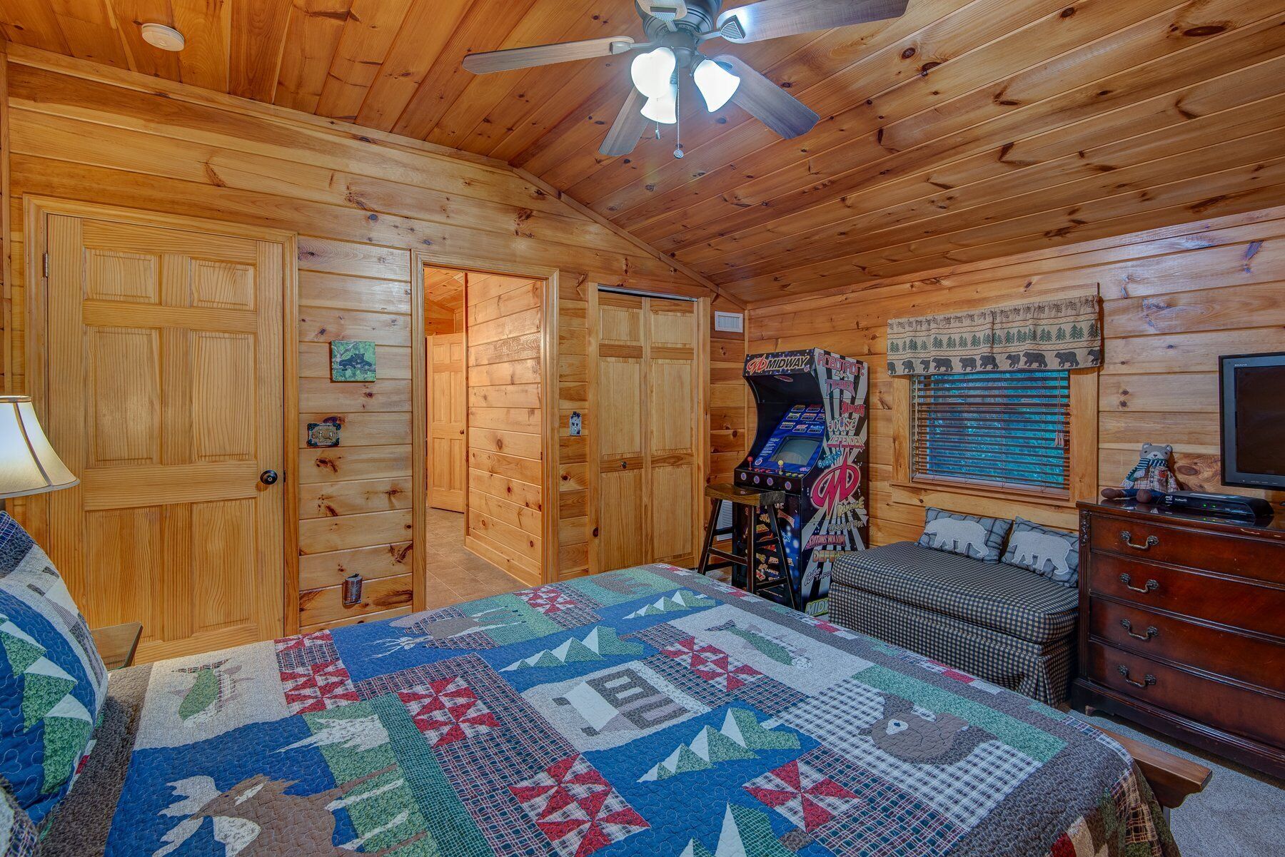 Une chambre confortable de style cabane, avec un plafond en bois, un ventilateur de plafond, un lit avec une courtepointe, une commode et une piste de voitures miniatures de la marque Midway.