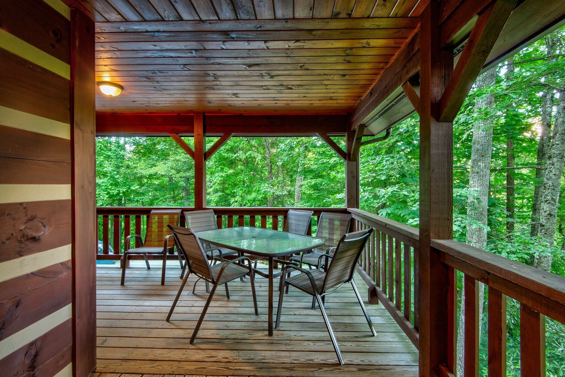 A covered outdoor deck with a glass table and chairs, surrounded by wooden railings and a view of a forested area.