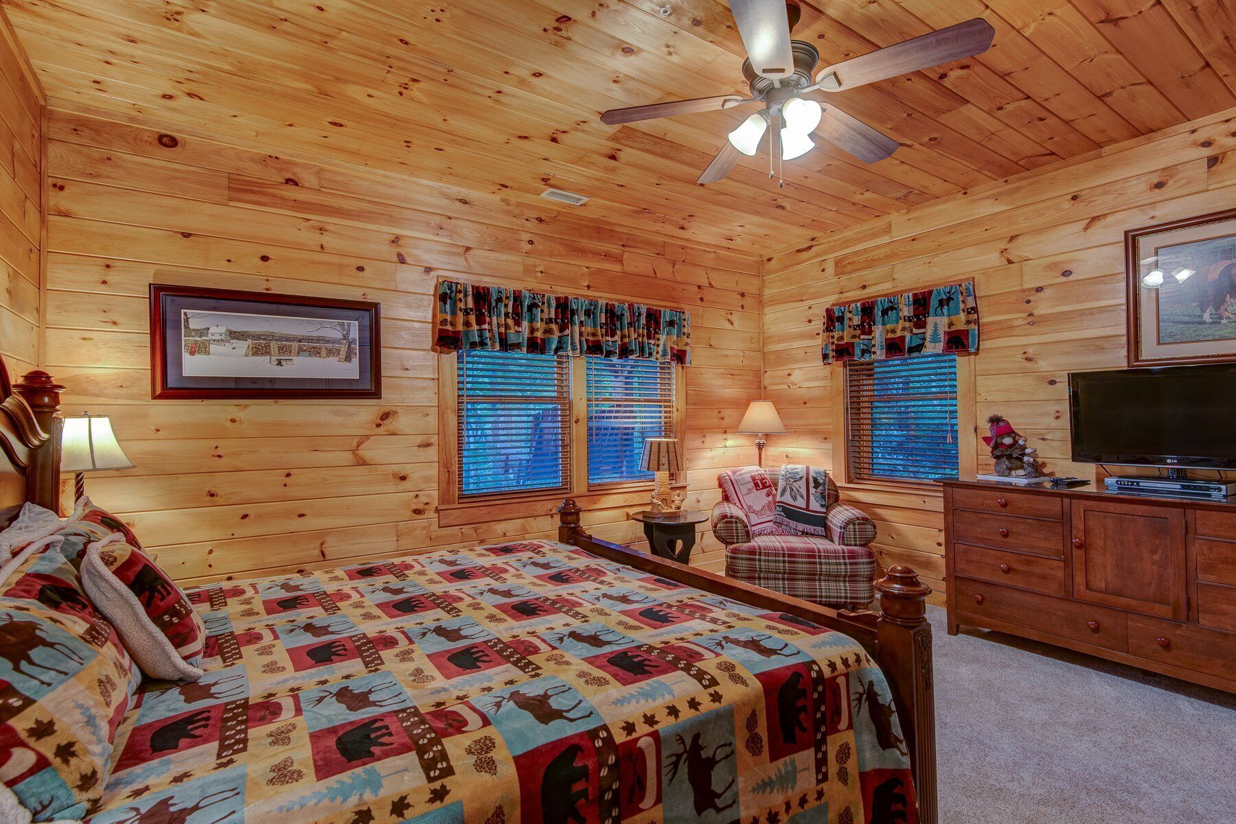 A cozy log cabin bedroom with a bed, a chair, a dresser, a ceiling fan, and a television.