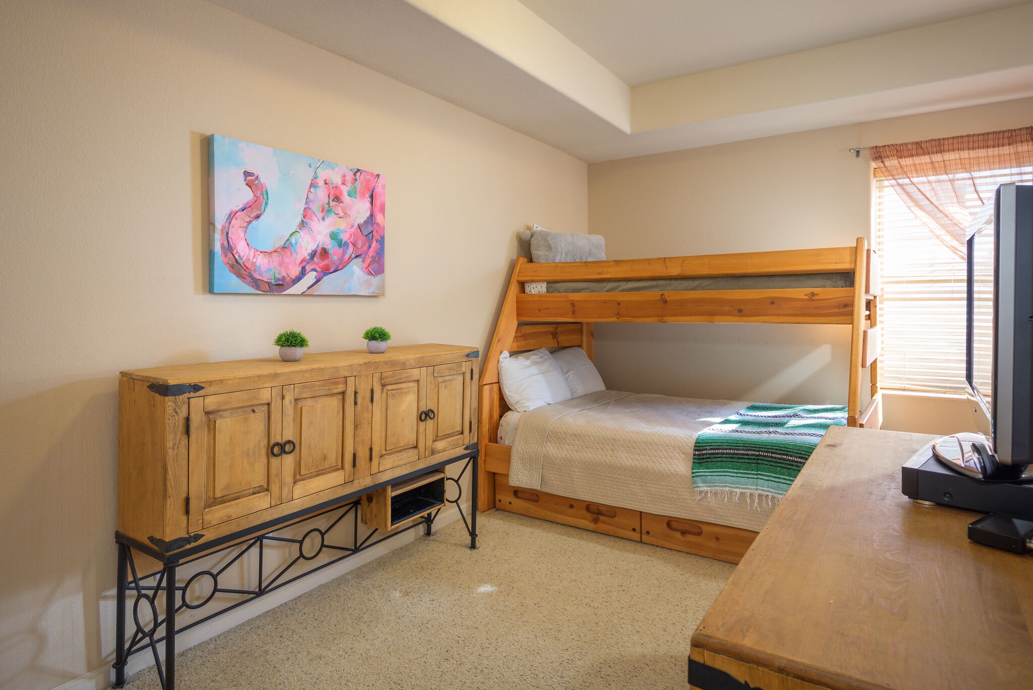 Una cama litera con cabecera de madera, un mueble pequeño con dos puertas, un televisor sobre un pedestal de madera y un cuadro en la pared.