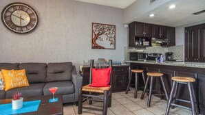 A modern kitchen with dark cabinetry, a microwave, and a coffee maker. There is a bar area with stools and a red chair. A round wall clock is visible above the kitchen counter.