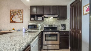 A kitchen with granite countertops, stainless steel appliances, dark wooden cabinets, and a decorative tree wall art piece.