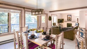 Zona pranzo con tavolo in legno apparecchiato per quattro persone, circondato da sedie in vimini con cuscini viola.