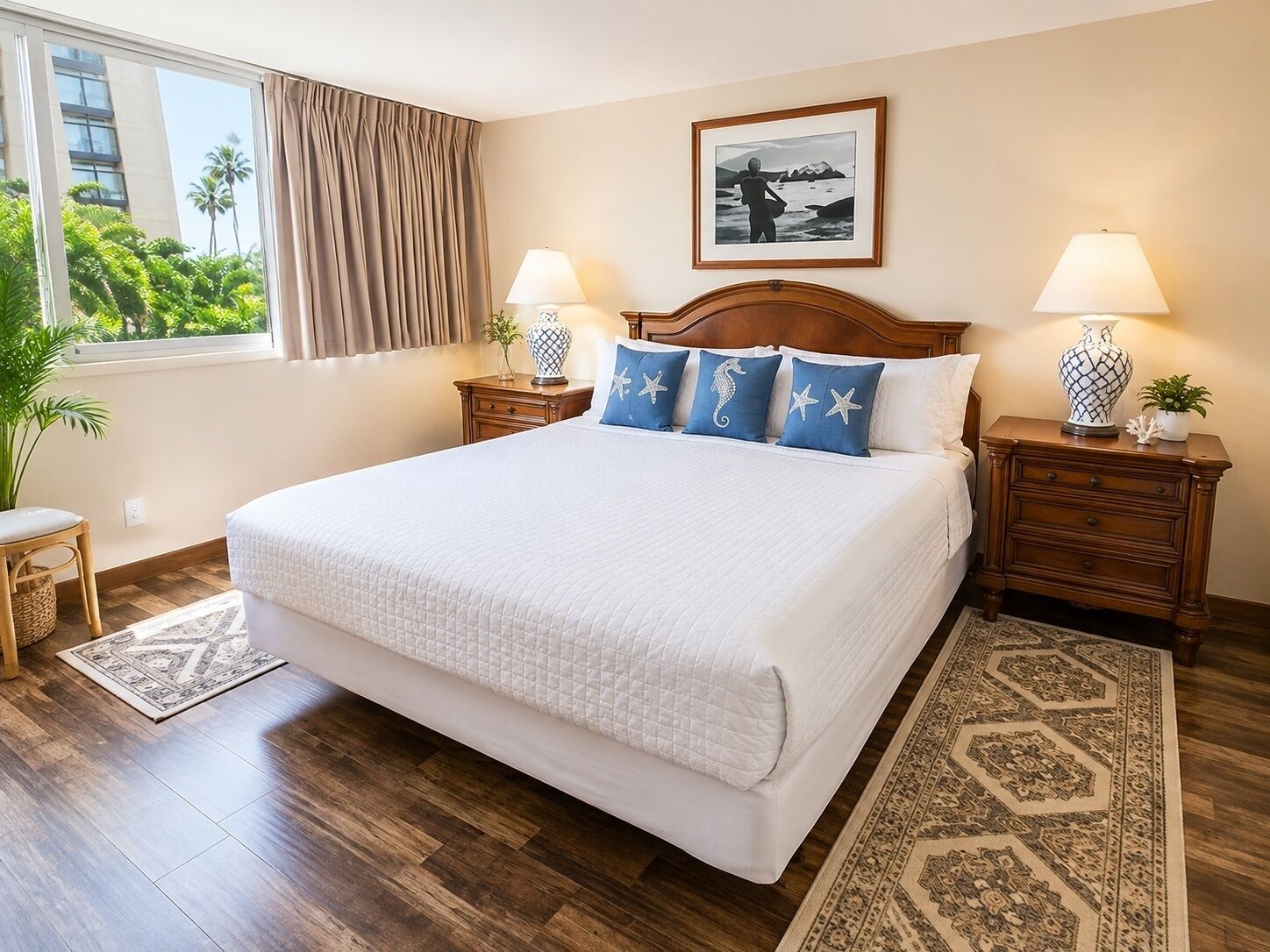 A hotel room with a large bed, wooden headboard, two bedside tables with lamps, a framed picture on the wall, and a view of palm trees through the window.