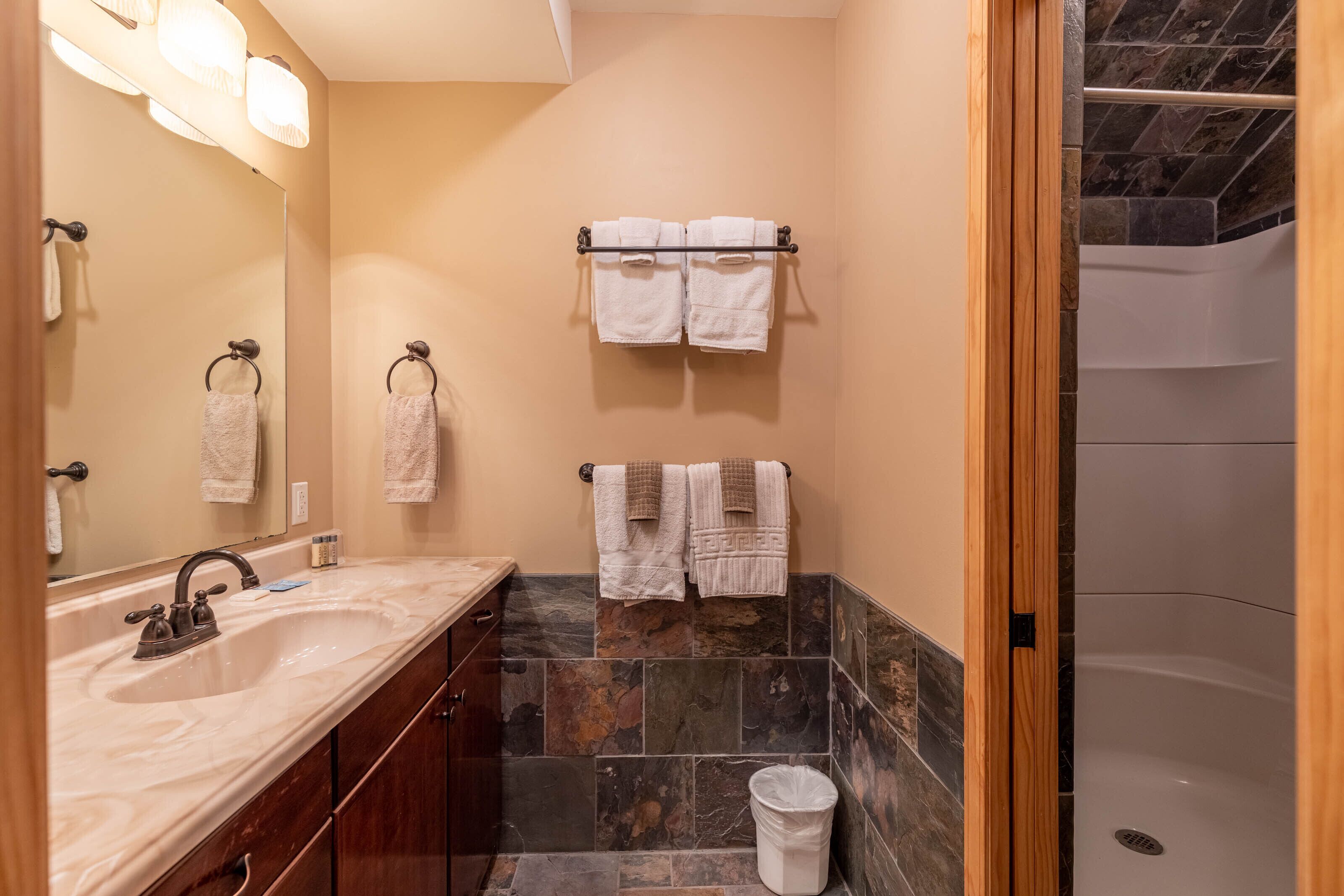 A bathroom with a marble sink, a mirror, a towel rack with multiple towels, a shower area, and a trash bin.