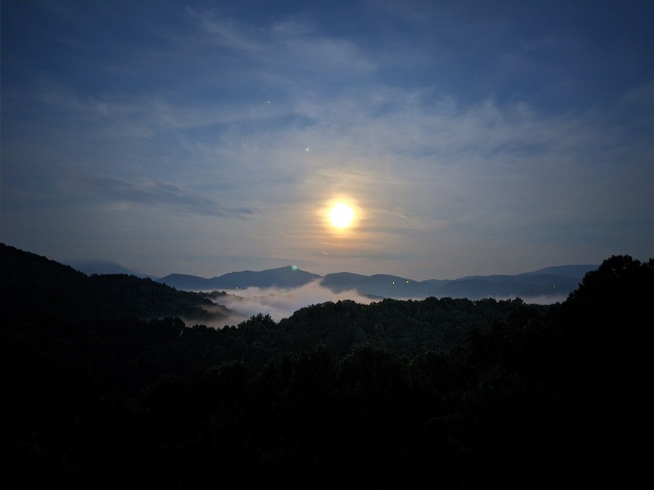 Eine Berglandschaft im Zwielicht, mit sichtbarer Sonne und weit entfernten Lichtern.