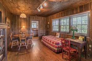 A cozy room with wooden walls and floors, a plaid bed, a wooden table with chairs, and a window with blinds.