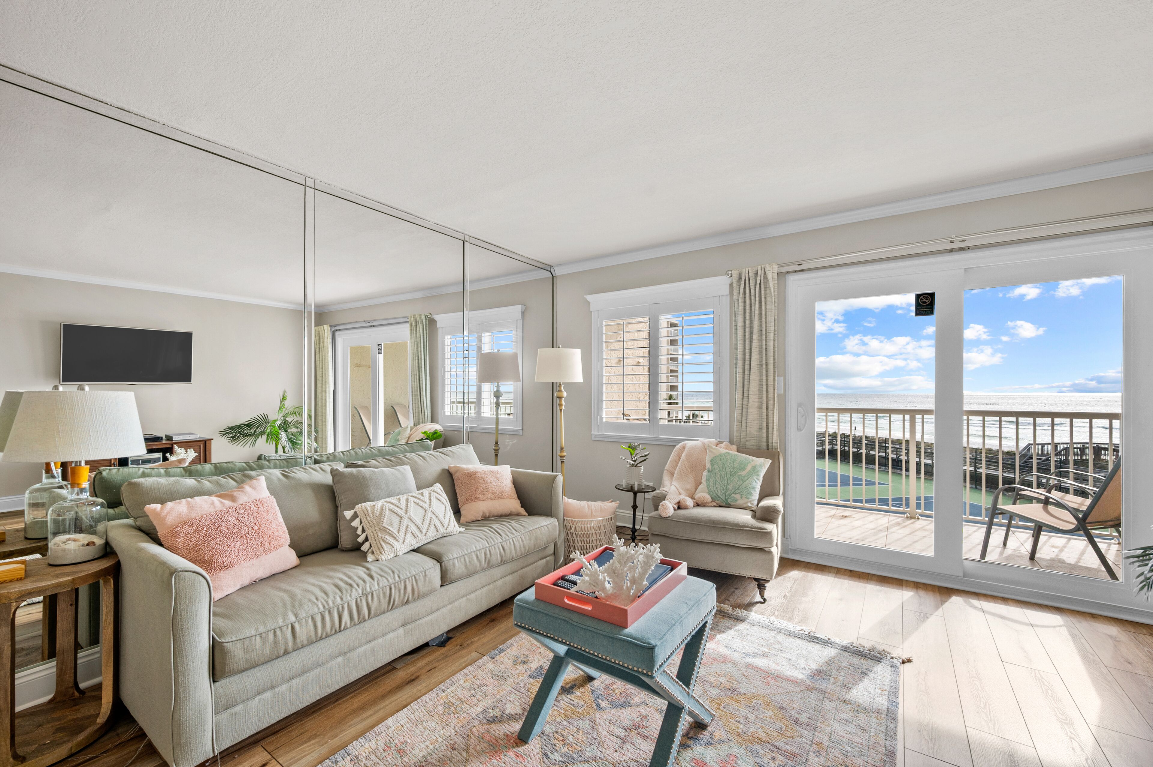 A spacious living room with a large window offering a view of the beach, a comfortable sofa, a coffee table, and a television on the wall.
