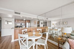 Une cuisine moderne avec des meubles blancs, un coin repas doté d’une table ronde et de chaises blanches, et un salon avec un canapé et une lampe.