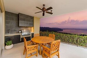 A balcony with a wooden table and chairs, a ceiling fan, and a view of a body of water at sunset.