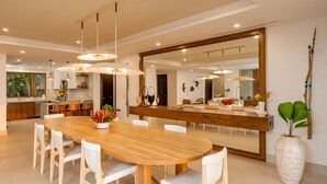 A modern kitchen with a wooden dining table, white chairs, pendant lights, and a large mirror.