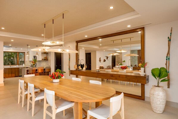 A modern kitchen with a wooden dining table, white chairs, pendant lights, and a large mirror.