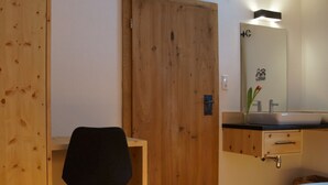 Une pièce avec une porte en bois, un bureau avec une chaise noire, un lit recouvert d’une couvre-lit à motifs floraux, une salle de bain avec un miroir, un lavabo et un porte-serviettes.