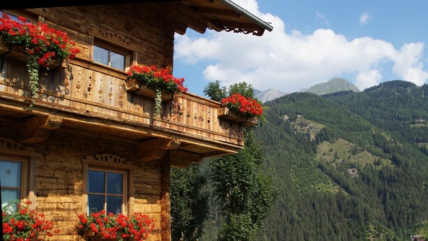 Un chalet en bois doté d’un balcon orné de fleurs rouges, situé dans un cadre montagneux.