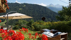 Une terrasse en bois avec un parasol beige, entourée de fleurs rouges et offrant une vue sur la montagne.