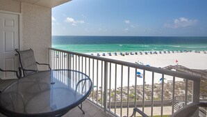 A balcony with a glass table and chairs, overlooking a beach with umbrellas and people.