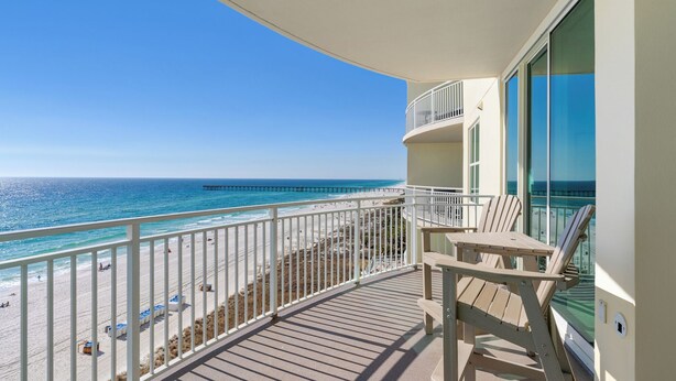 Un balcon offrant une vue sur la plage et l’océan, avec une terrasse en bois, une balustrade blanche et une table entourée de chaises.