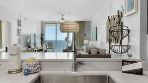 Une cuisine moderne dotée d’un évier en acier inoxydable, d’un luminaire suspendu en osier et offrant une vue sur l’océan grâce à une porte-fenêtre coulissante.