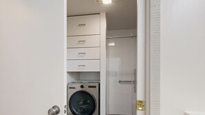 A laundry room with a front-loading washing machine, a glass shower enclosure, and a white tiled floor.
