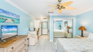 Un dormitorio con cama, televisor, cómoda, ventilador de techo y puerta que da a otra habitación.