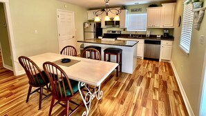 A modern kitchen with white cabinets, a central island, and a dining area with wooden chairs. - MI CASA DEL MAR - So close to the beach, perfect house for 4! (Caplen)