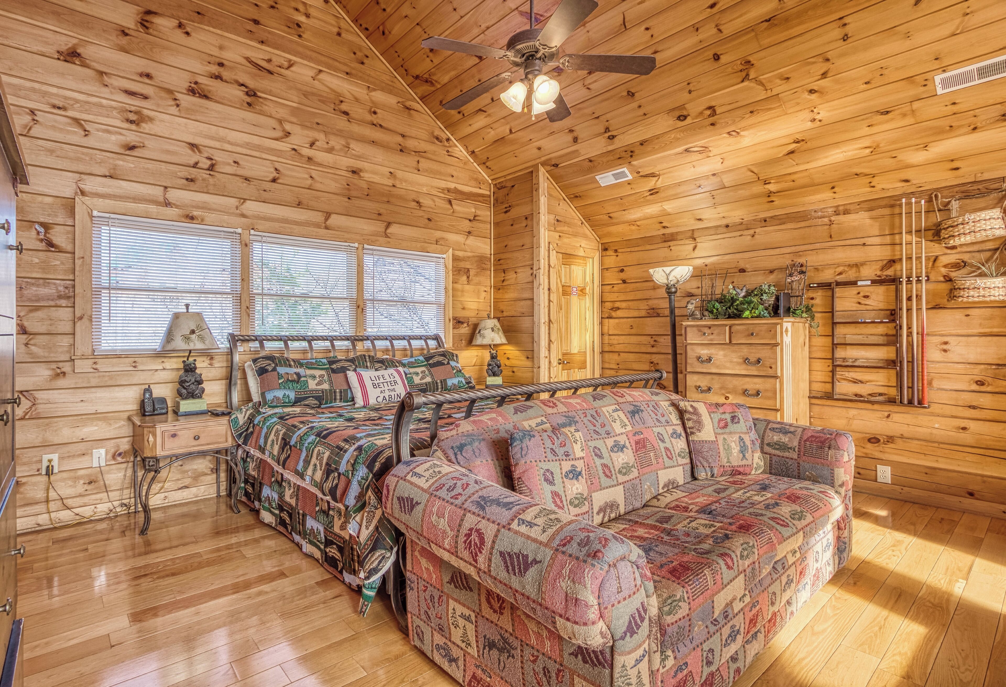 A cabin interior with a wooden floor, a bed with a colorful quilt, a sofa, a ceiling fan, and a window with blinds.