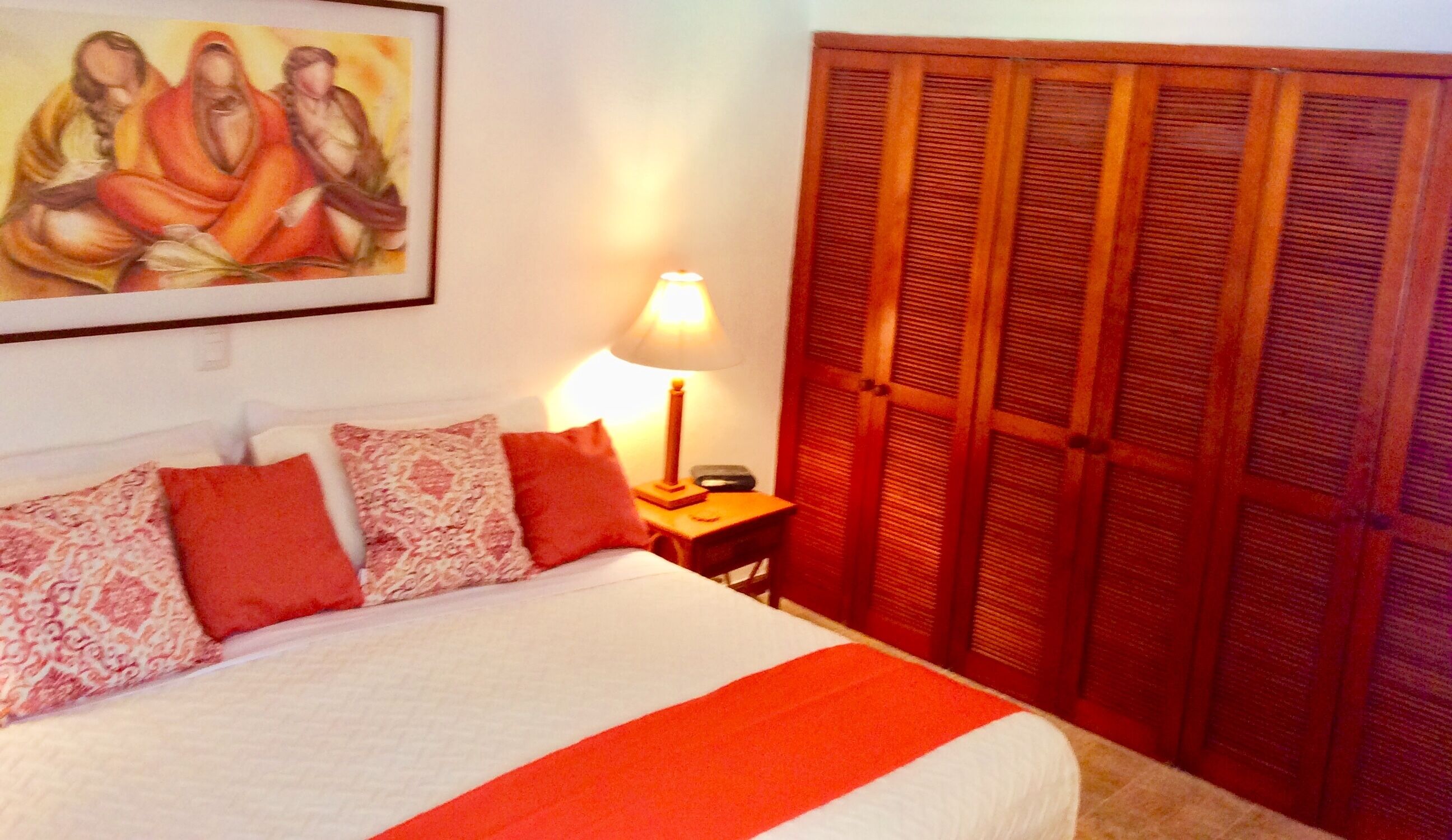 A bedroom with a bed, pillows, a nightstand, a lamp, a painting, and a wooden wardrobe.