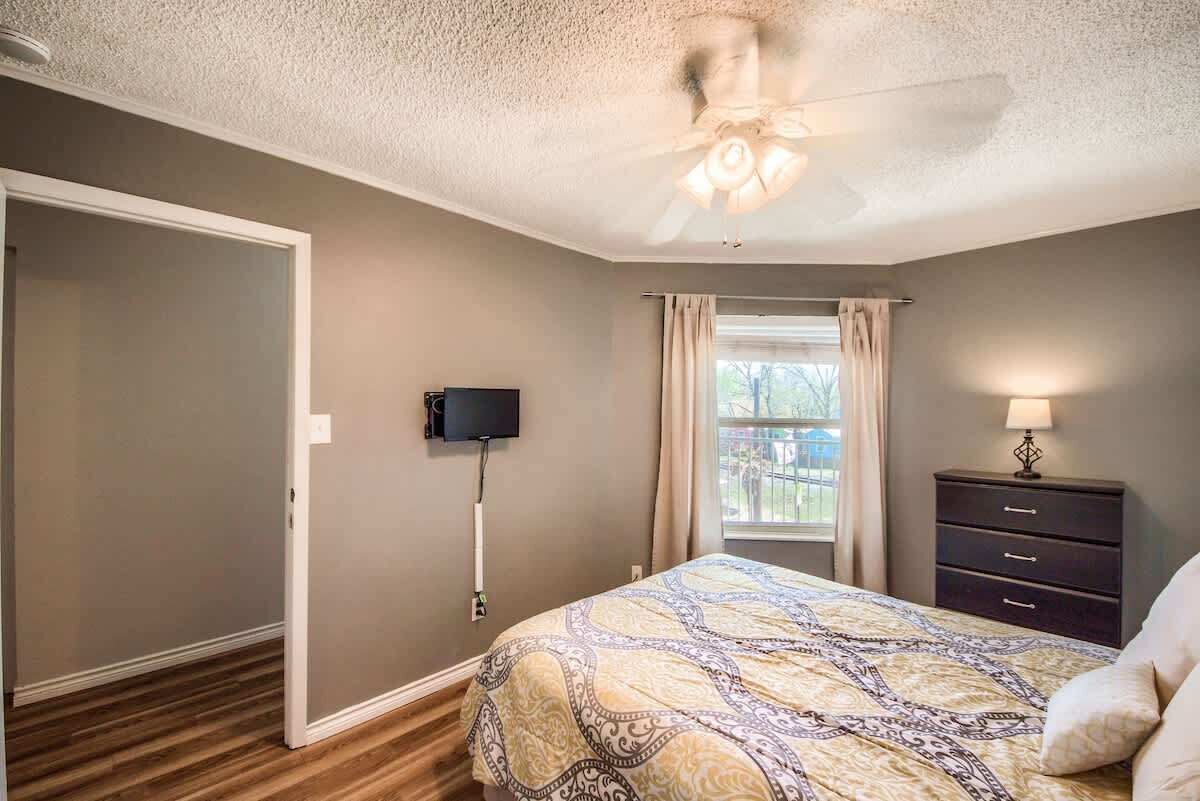 A bedroom with a bed, a ceiling fan, a TV, a window with curtains, and a nightstand with a lamp.