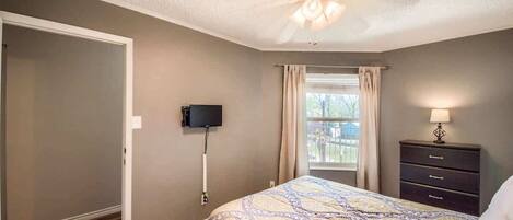 A bedroom with a bed, a ceiling fan, a TV, a window with curtains, and a nightstand with a lamp.