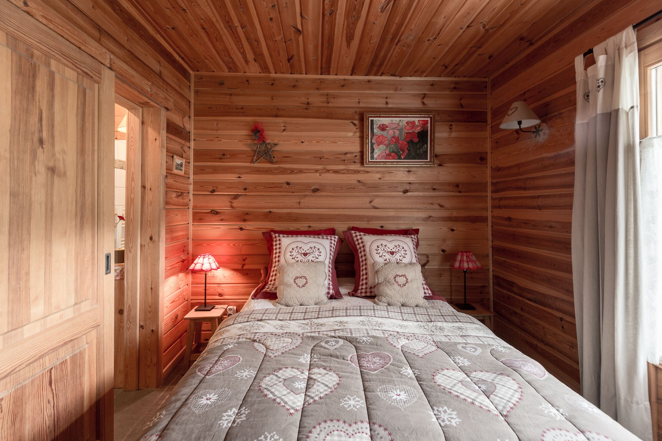 Un dormitorio de cabaña de madera con una cama, dos lámparas de noche, una mesita de noche, un cuadro enmarco y una ventana con cortinas.