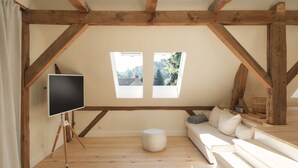 Un dormitorio tipo loft con techo de madera, un ventanal grande, un televisor sobre un trípode, un sofá y un reposapiés redondo pequeño.
