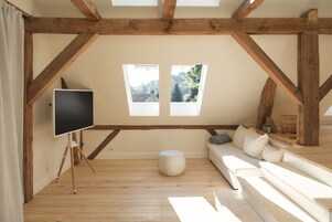 Un dormitorio tipo loft con techo de madera, un ventanal grande, un televisor sobre un trípode, un sofá y un reposapiés redondo pequeño.