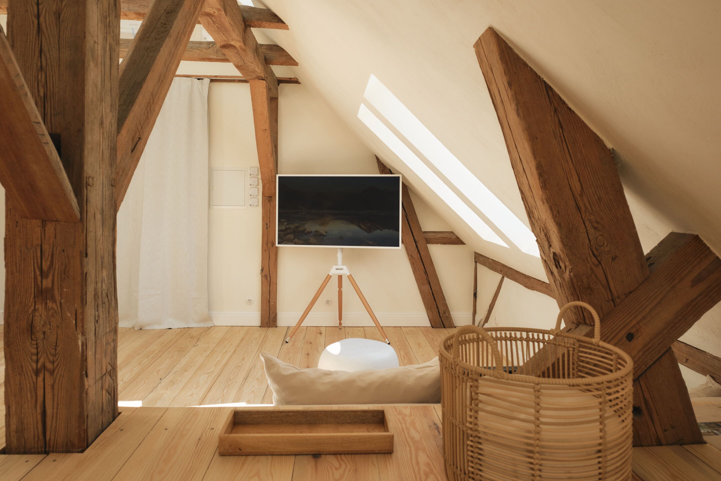 A cozy attic room with wooden beams, a white cushioned bench, a wicker basket, and a mounted TV on a tripod.