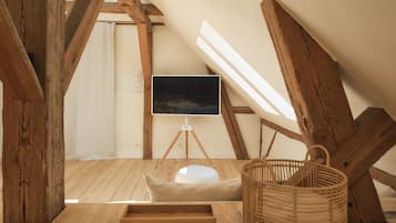 A cozy attic room with wooden beams, a white cushioned bench, a wicker basket, and a mounted TV on a tripod.