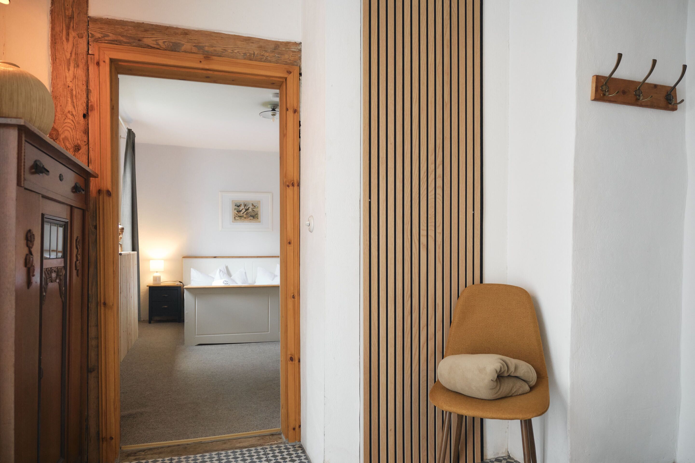 Een kamer met een houten deur, een stoel, een kapstok en een bed met nachtkastje.