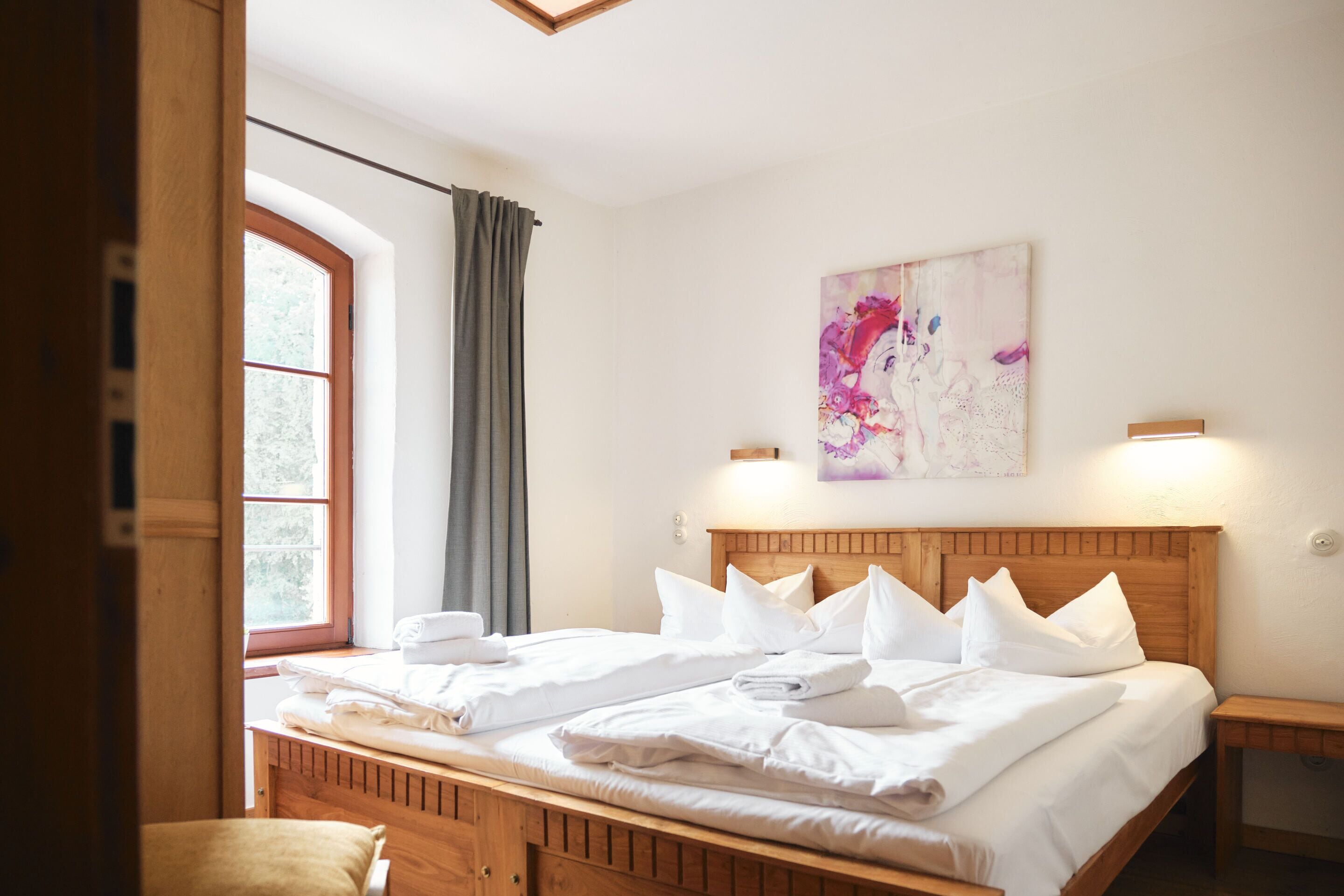 Una cama bien hecha con sábanas y almohadas blancas, un cabecero de madera, un cuadro en la pared y una ventana con cortinas.