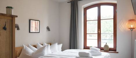 A neatly made bed with white linens and pillows, a wooden headboard, a window with curtains, a wall-mounted lamp, and a small potted plant on the shelf.