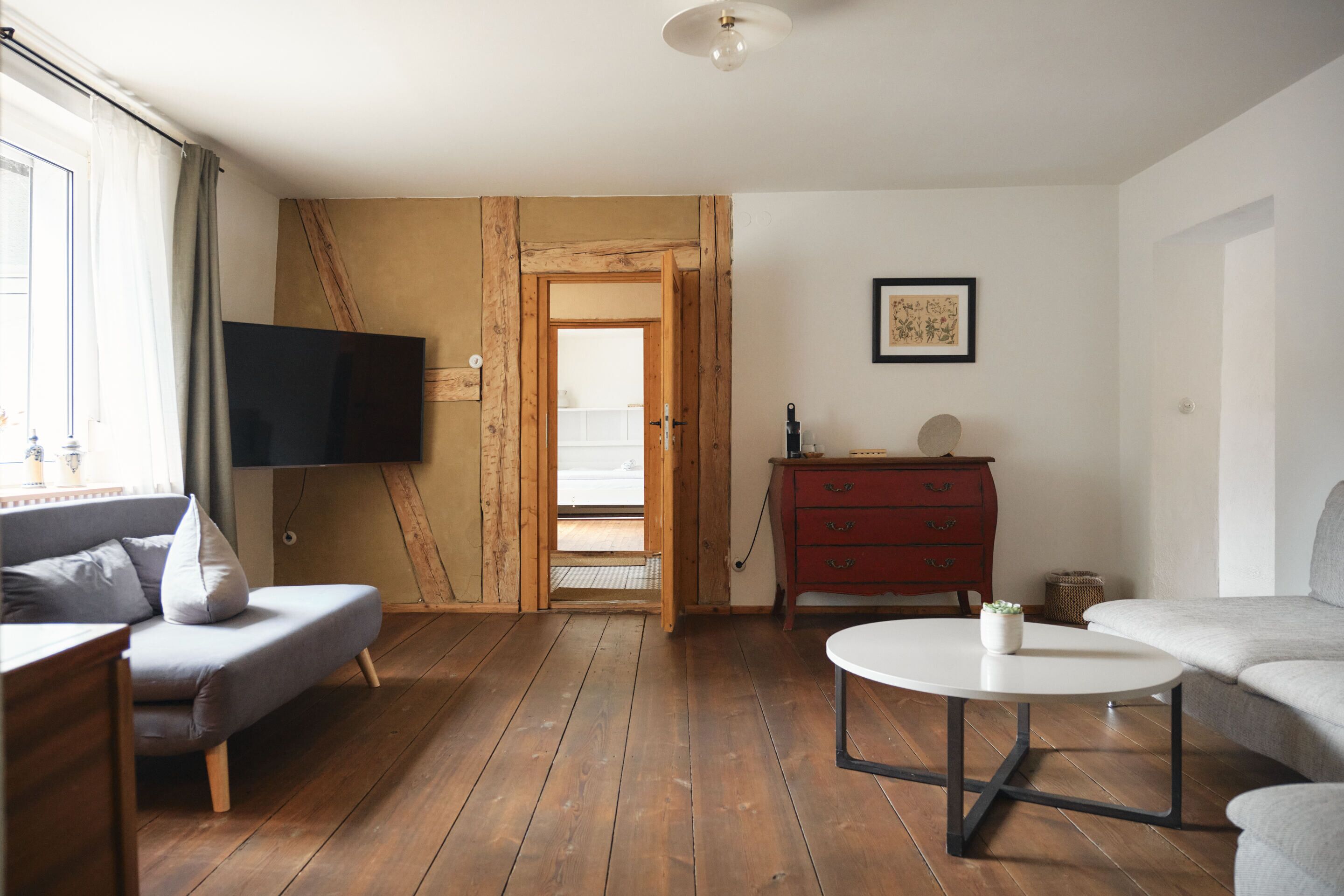 A living room with a wooden floor, a grey sofa, a red dresser, a white coffee table, and a TV mounted on the wall.