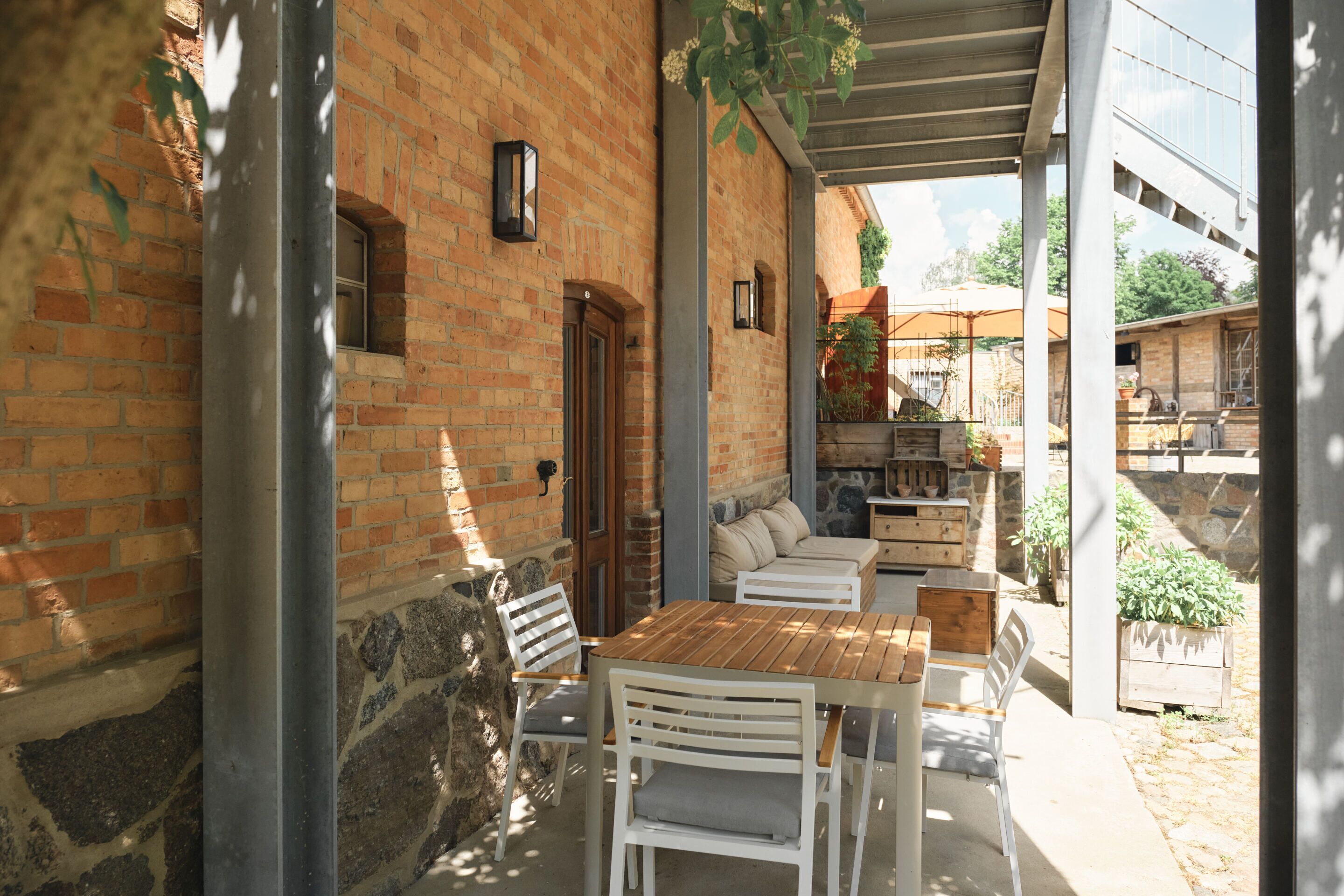 An outdoor patio with a wooden table and white chairs, a brick wall, and a covered area with a view of the surrounding landscape.